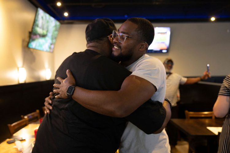 Two men share a warm hug at the Civic Flint gathering, one smiling in glasses and a white T-shirt, while others mingle in the background of the restaurant.