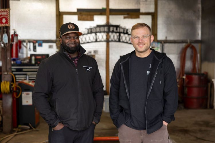 Aaron Dunigan and Matthew Soulia stand smiling together in a metal workshop, with a decorative arch reading "Forge Flint" and various tools and equipment in the background.