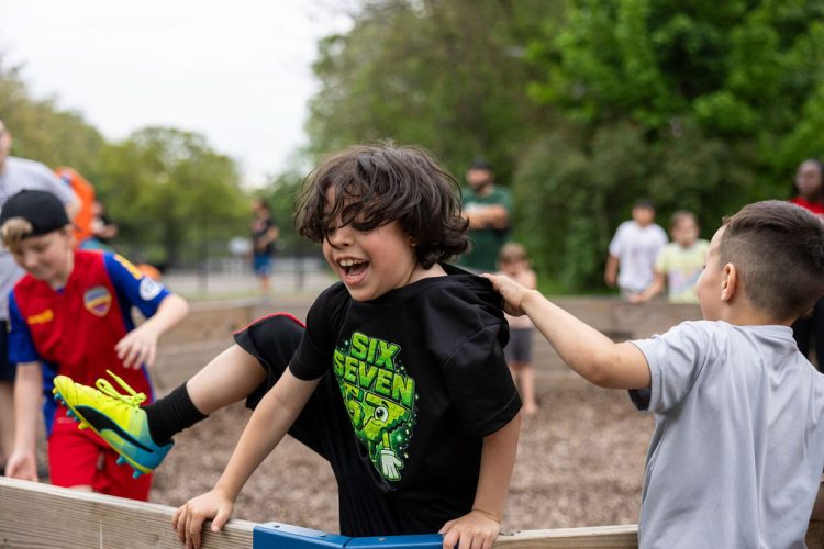A boy laughs joyfully while climbing over the wooden wall of a gaga ball pit, with another child pulling at his shirt and kids playing in the background.