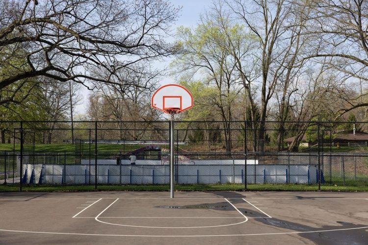 An empty outdoor basketball court with an orange-and-white hoop, enclosed by a chain-link fence, with bare trees and a fenced-in area visible in the background at Mott Park.