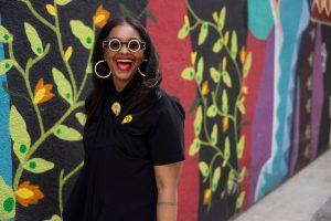 A woman is laughing in front of a colorful mural. She is wearing a black shirt with glasses.