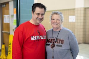 A man and woman pose for a photo. The man is wearing a red long sleeve shirt with Grand Blanc Bobcats on it. The woman is wearing a grey long sleeve shirt that also says Grand Blanc bobcats.