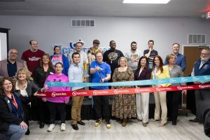 A group of people stand in front of ribbons at riibbon cutting. A man holds a giant pair of scissors.