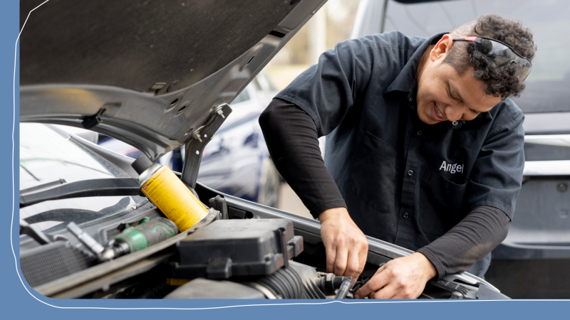 A mechanic named Angel, wearing a black work shirt and safety glasses pushed up on his head, smiles while working under the open hood of a car.