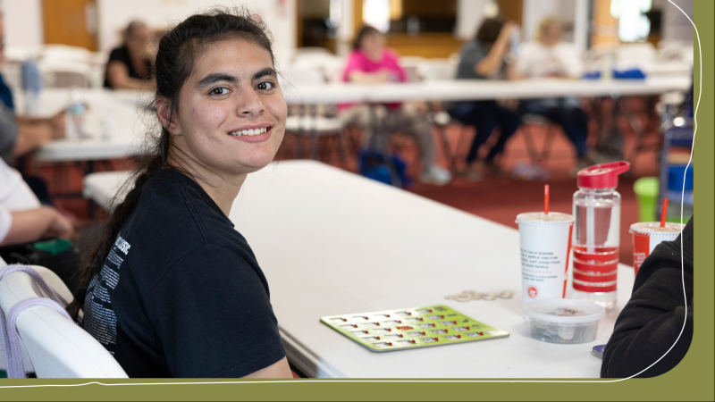 A girl with brown hair is seated and smiling at a table with a bingo card. She is attending an Xceptional Heroes bingo night event.