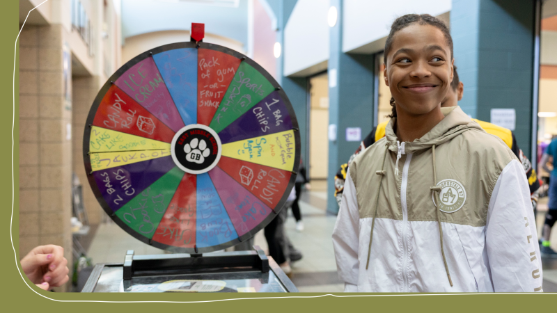 A boy, wearing a zip up jacket, is grinning next to a spinning wheel.