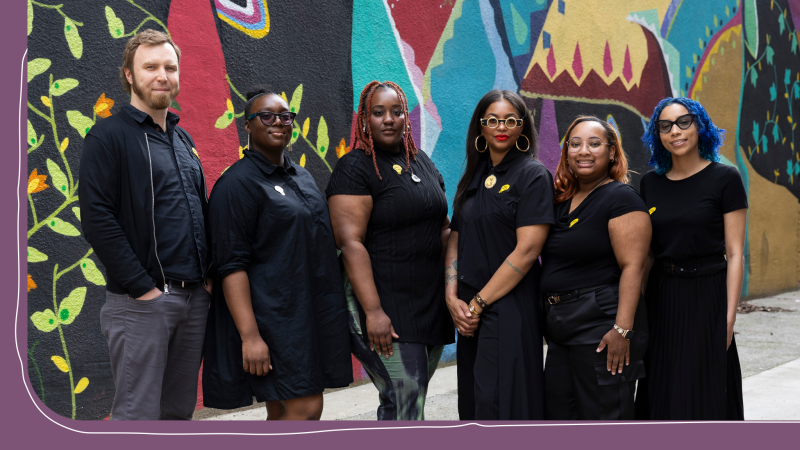 Six individuals pose for a photo in front of a colorful mural.