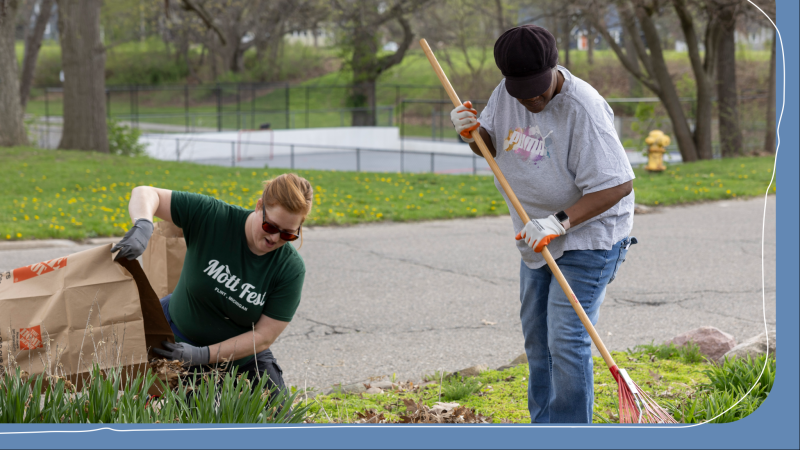 Two volunteers clean up a garden bed in Mott Park, one holding open a paper yard-waste bag while the other rakes leaves with a wooden-handled rake near a fenced tennis court.