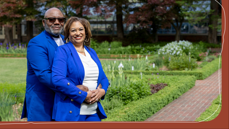 Wendell and Sheryl Thompson pose together smiling in matching blue blazers in a formal garden, with manicured hedges and trees in the background.