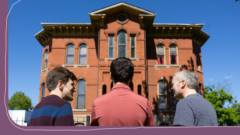 Three people stood outside an ornate red-brick building with arched windows, viewed from behind, looking up at the historic architecture under a clear blue sky.
