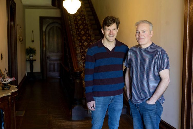Brian Tesler and William Gainey stand smiling side by side in the wood-paneled front hallway of a historic home, with an ornate staircase and antique furnishings behind them.