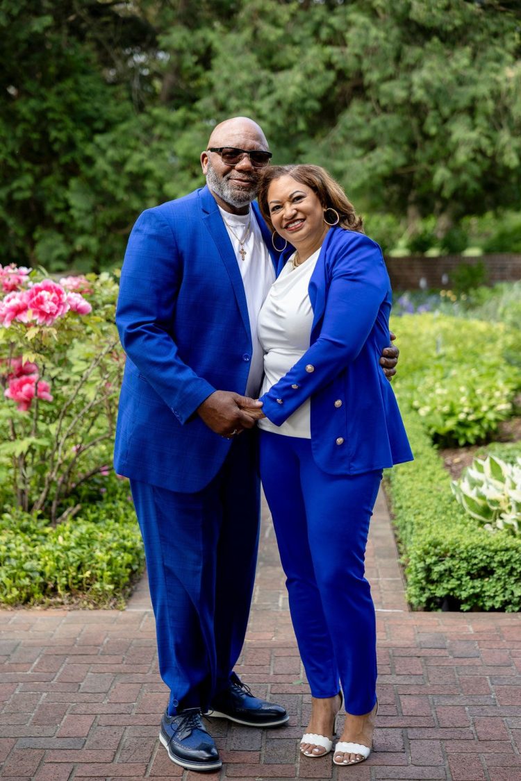 Wendell and Sheryl Thompson pose together smiling in matching blue blazers in a formal garden, with manicured hedges and trees in the background.