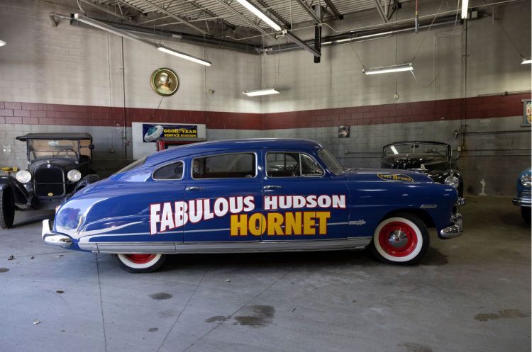 A vintage dark blue Hudson Hornet race car with red wheels and white-lettered "FABULOUS HUDSON HORNET" livery on its side, parked in a garage alongside other classic and antique cars.