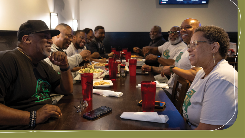 Members of Civic Flint sit together at a long table at Paul's Steakhouse, laughing and talking over a shared meal, several wearing matching green-and-white "Flint" T-shirts.
