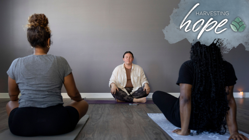 A yoga instructor and two participants sit on floor mats and engage in meditation.